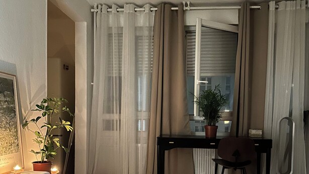 A bedroom with a bed, a desk, and a chair. There is a window with curtains, a potted plant, and a lamp on the desk.
