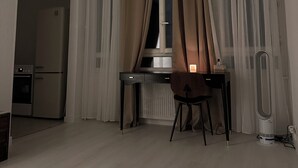 A modern, well-lit room with a dining table, chairs, a standing heater, and a kitchen area with a refrigerator and oven.