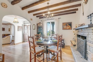 A dining room with a round table set for six, wooden chairs, a chandelier, a fireplace, and a view through a window. - Casa Naima (Alto Malcantone)