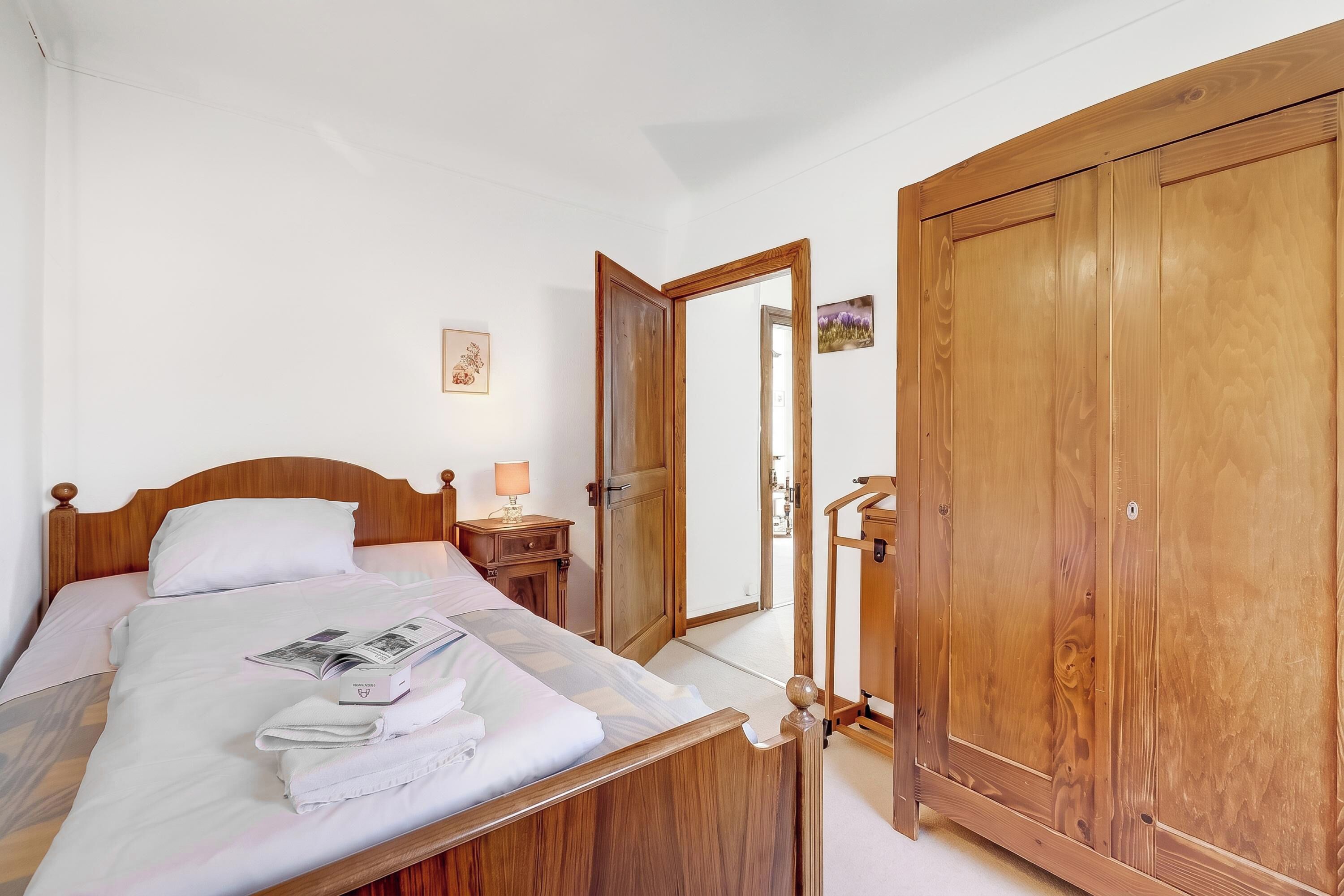 A hotel room with a wooden bed, a nightstand with a lamp, a wardrobe, and a mirror reflecting the room.