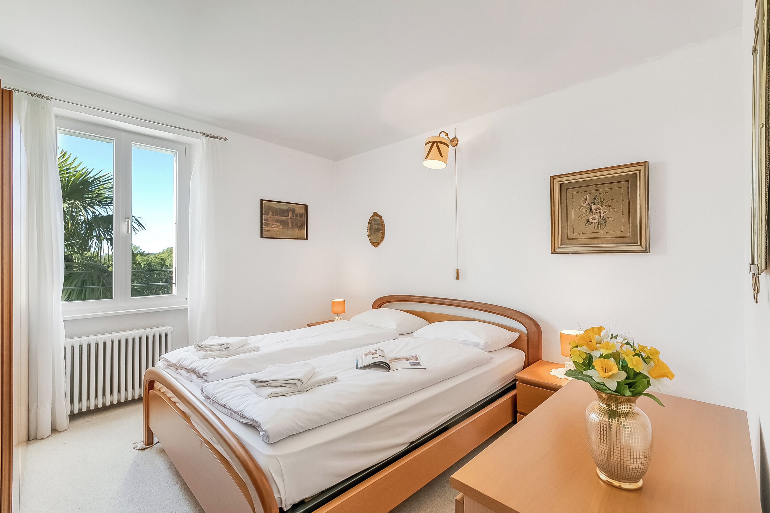 Un dormitorio con una cama, una mesita de noche, una lámpara, un florero con flores y dos cuadros en la pared.