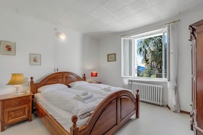 A bedroom with a wooden bed, bedside tables, a window with a view, and a radiator. - Casa Naima (Alto Malcantone)
