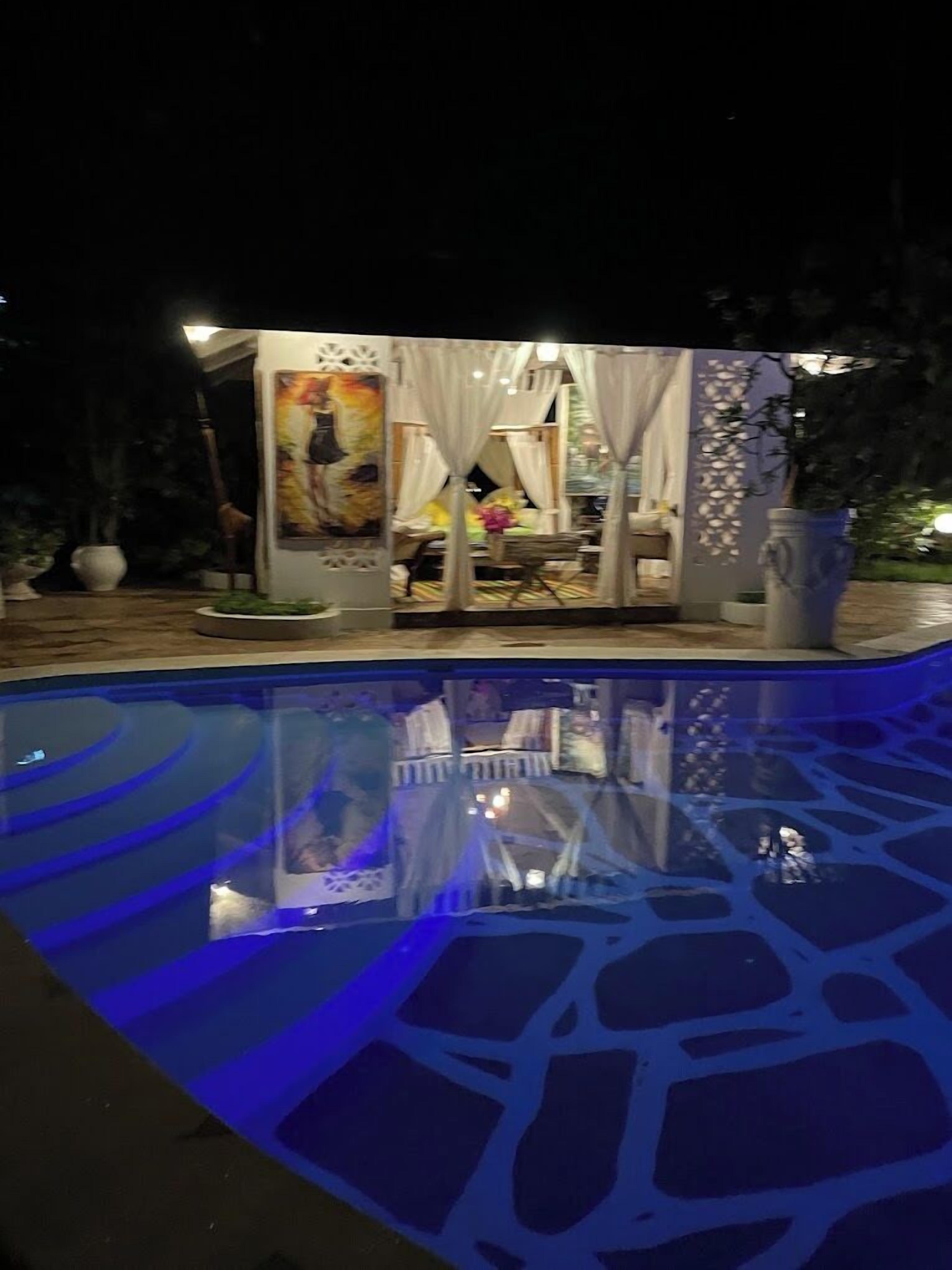 A nighttime view of a pool area with a glass enclosure, a mural, and a seating area with white curtains.