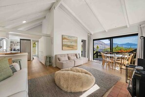 A spacious living area with a large window, a sofa, a dining table, and a kitchen area. - Lake & Mountain Views | 2 Living Areas | Sleeps 8 (Wānaka)