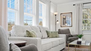 Ein modernes Wohnzimmer mit grauem Sofa, Couchtisch und grauem Sessel. Es gibt ein großes Fenster mit Jalousien, eine Stehlampe und ein gerahmtes Bild an der Wand.