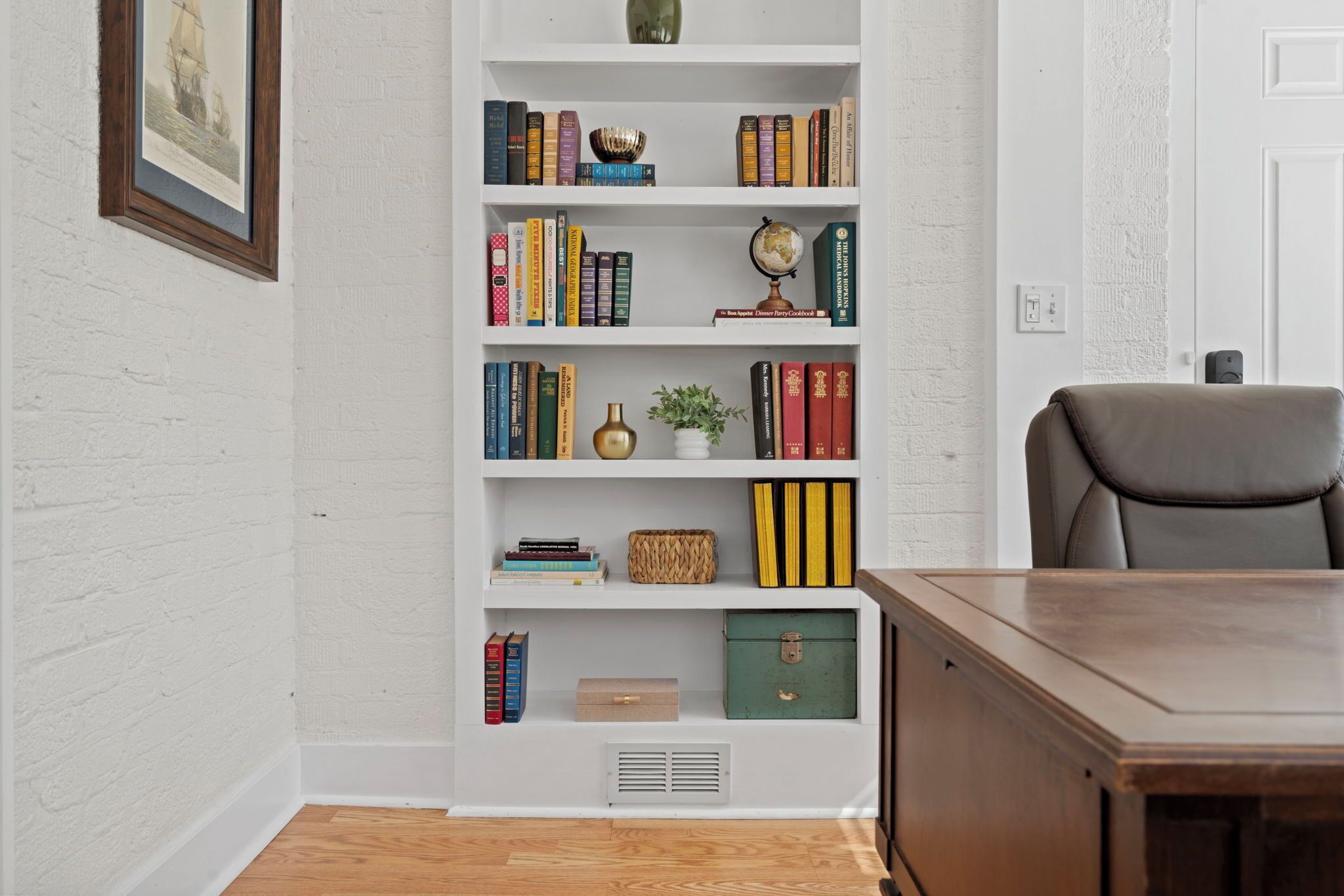 A room with a wooden desk, a bookshelf with various books and decorative items, a globe, and a framed picture on the wall.