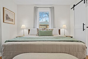 Ein Schlafzimmer mit einem großen Bett, zwei Nachttischlampen, einem Fenster mit Vorhängen und einem gerahmten Bild an der Wand.