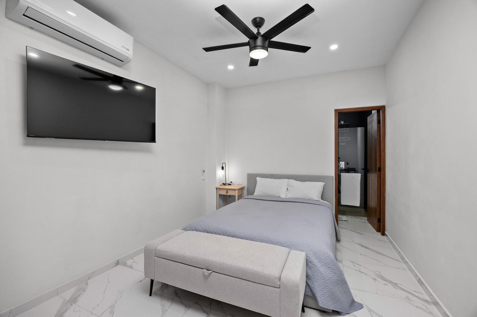 Een moderne slaapkamer met een bed, een nachtkastje, een flatscreen-tv, een airconditioner en een plafondventilator.