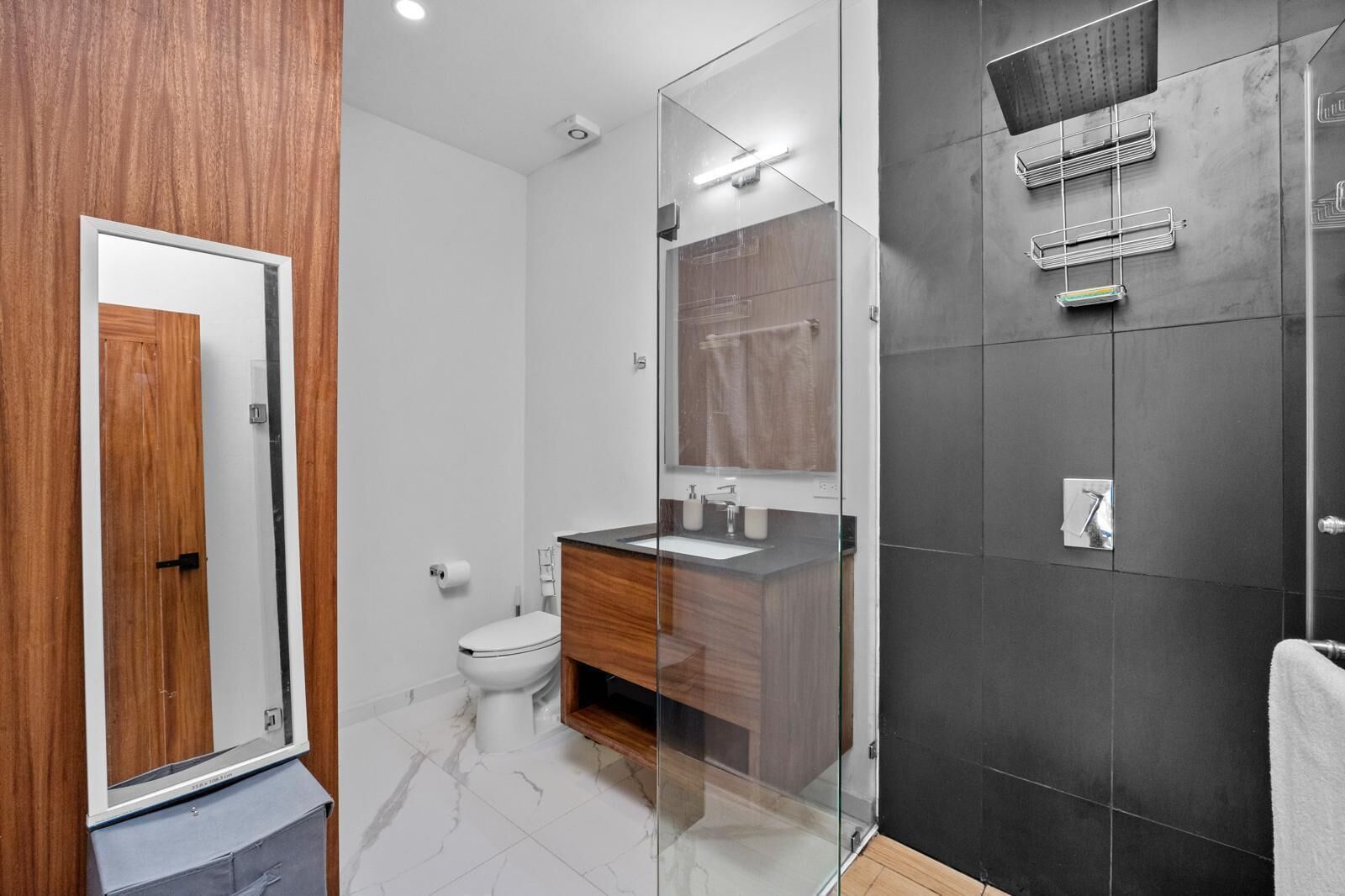 A modern bathroom with a wooden vanity, a glass shower enclosure, and a dark tiled shower area.