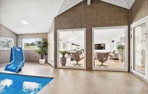 A modern indoor pool area with a blue slide, large windows, and a dining area visible through glass doors. - Amazing home in Idestrup with sauna (Idestrup)