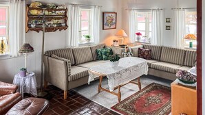 Un salon chaleureux avec un canapé à rayures, une petite table recouverte d’une nappe en dentelle, un lampadaire et une étagère décorative où sont exposées des pièces de poterie.