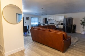 Ein modernes Wohnzimmer mit einer Ledersofa, einem Essbereich mit Tisch und Stühlen, einer Küche mit weißen Schränken und Geräten, sowie einem großen Spiegel an der Wand.