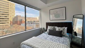 Une chambre à coucher moderne avec une grande fenêtre, un lit avec une literie à motifs, une tête de lit, une table de chevet et une œuvre d’art encadrée accrochée au mur.