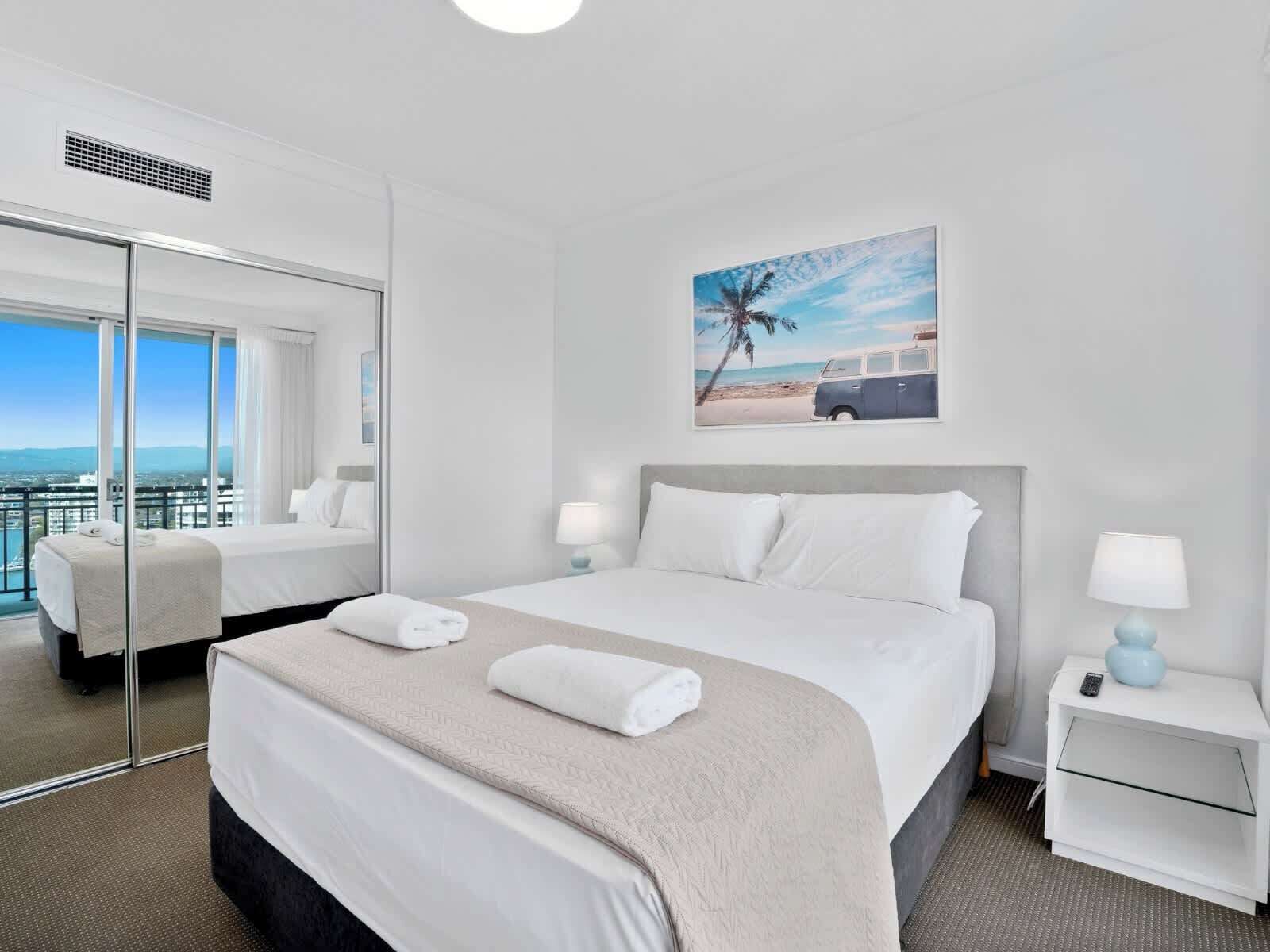 Ein Hotelzimmer mit einem großen Bett, zwei Nachttischen mit Lampen, einem großen Spiegel und einem Bild mit einer Strandansicht.