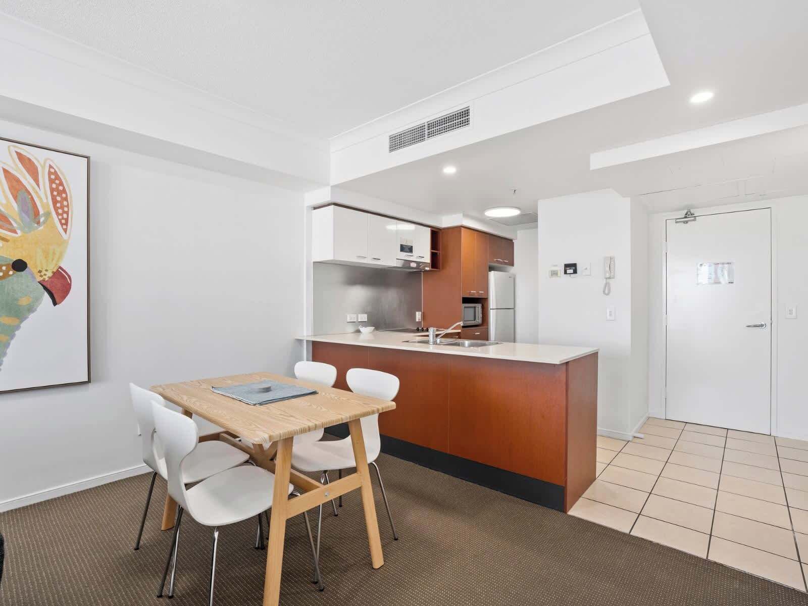 A modern kitchen with a central island, white cabinets, and stainless steel appliances. There is a dining area with a wooden table and white chairs.