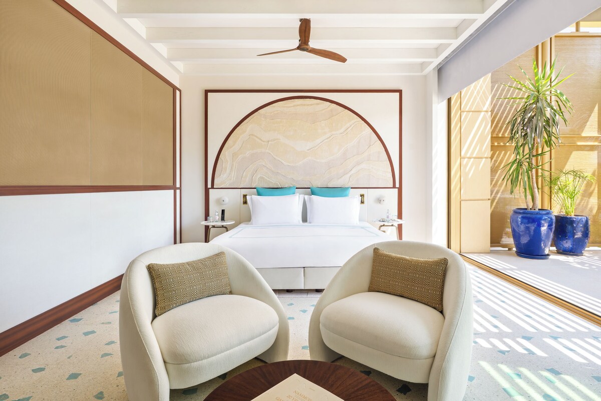 Habitación de hotel con una cama, dos sillones, un ventilador de techo y un ventanal con plantas en macetas.