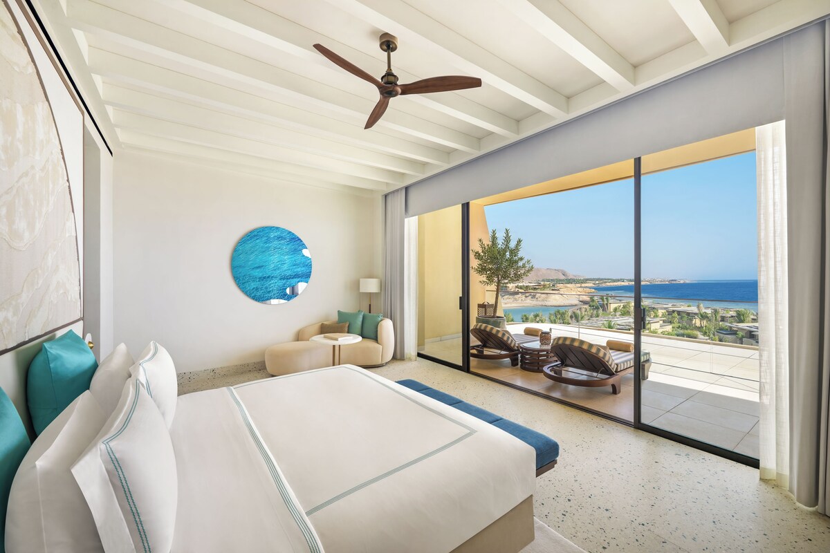 Habitación de hotel con cama, un adorno azul circular en la pared, ventilador de techo y balcón con vista al mar.
