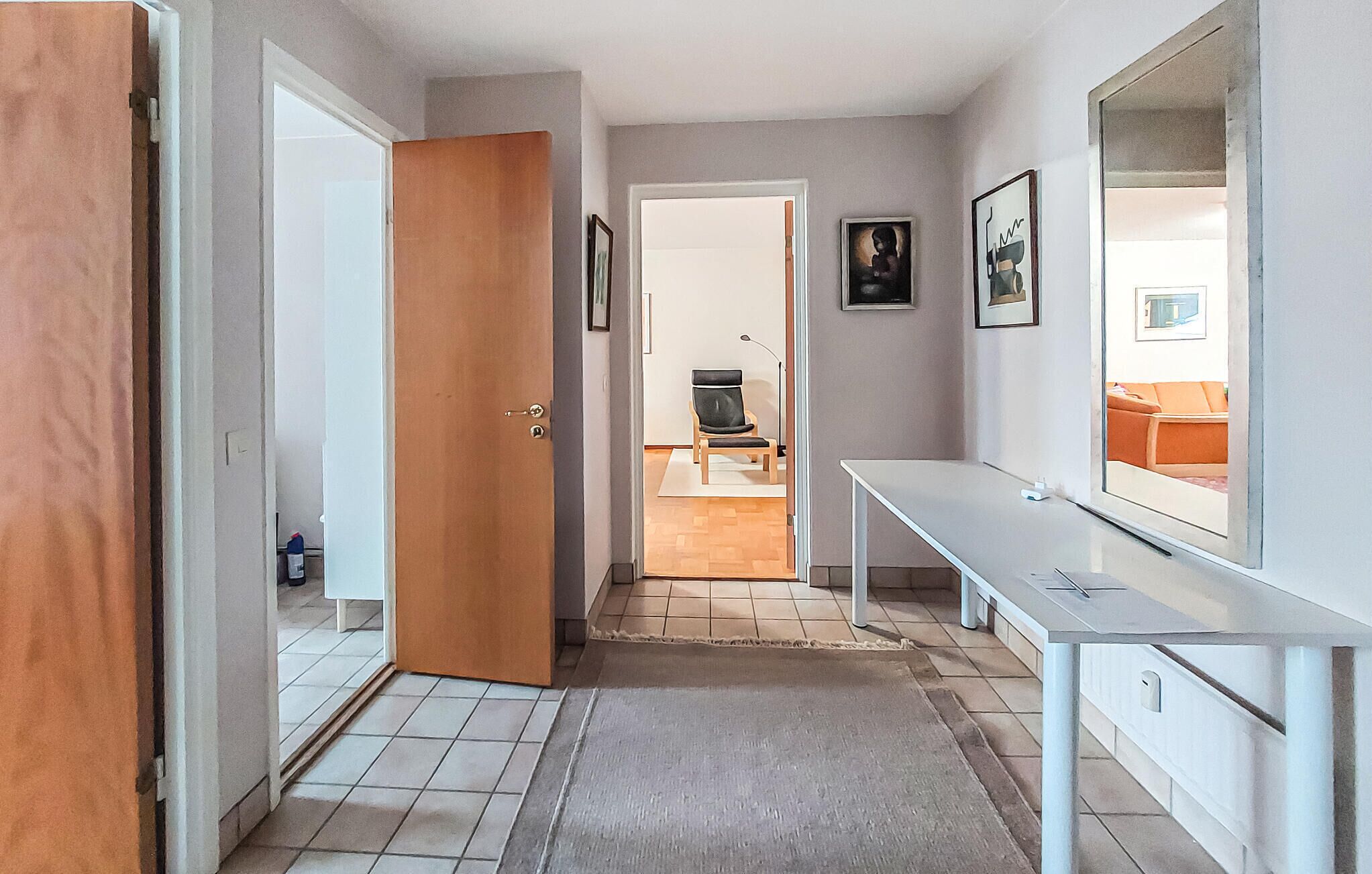 A hallway with a wooden door, a white desk, and a chair. There is a framed picture on the wall and a view into another room with a sofa.