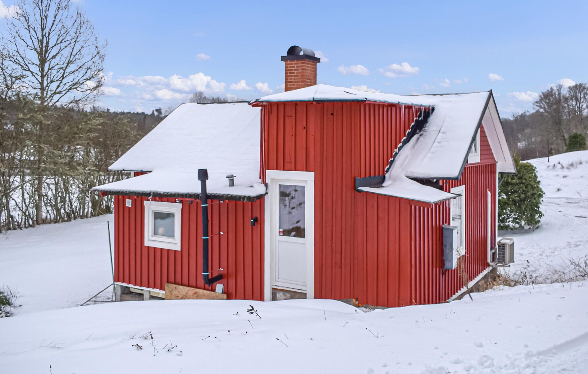 A red wooden house with a snow-covered roof, a chimney, and a small porch area.