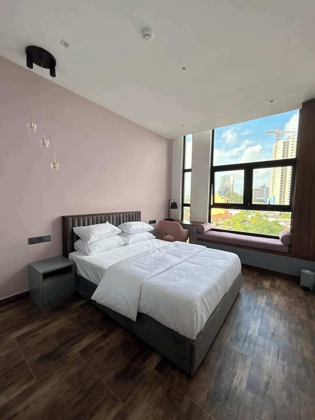 A modern bedroom with a large bed, a nightstand, a bench by the window, and a view of buildings outside.