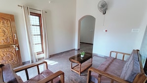 Un salon comprenant un canapé, deux fauteuils, une petite table avec une plante, une porte en bois et un ventilateur fixé au mur.