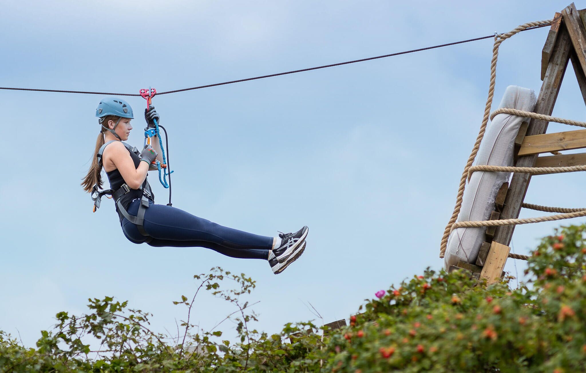 A person zip-lining with safety gear, including a helmet and harness, over a wooden structure with ropes.