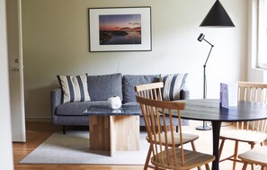 A modern living room with a grey sofa, wooden chairs, a round table, and a framed picture on the wall.