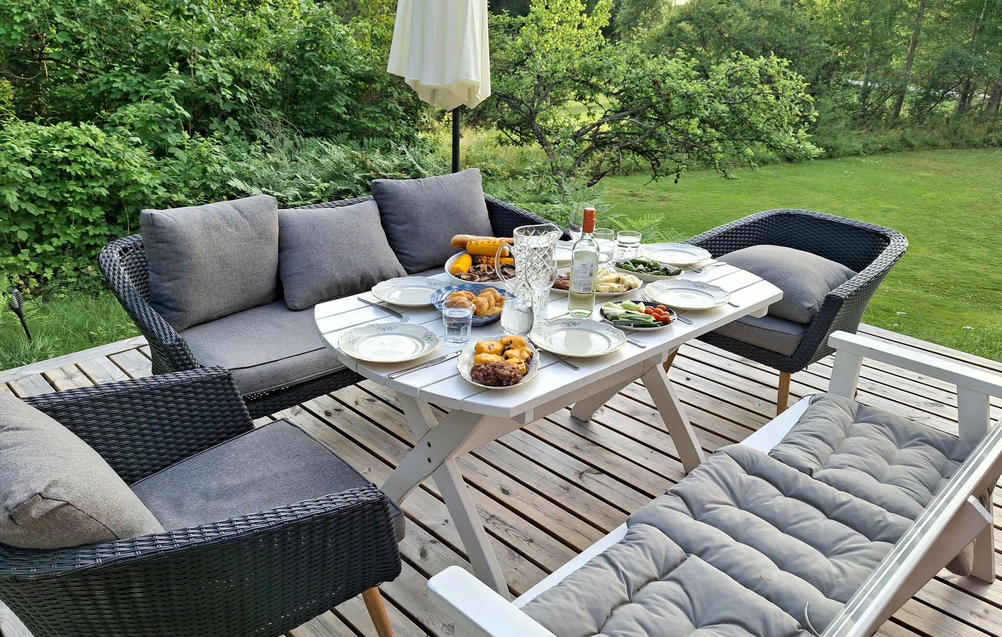 An outdoor dining area with a table set for four, surrounded by wicker furniture and a lush green backdrop.