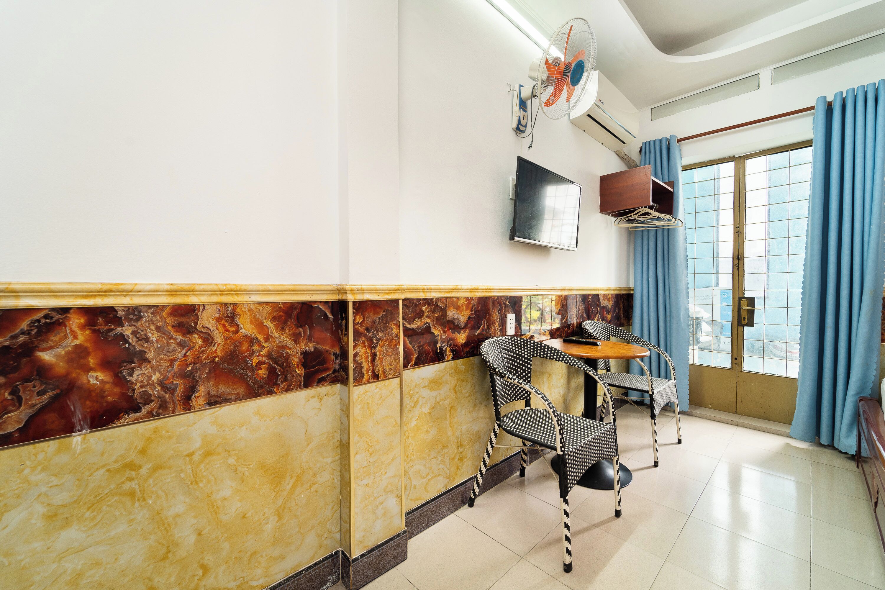 A hotel room with a marble wall, a TV mounted on the wall, a table with chairs, and a window with curtains.
