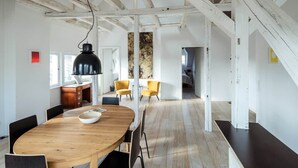 Um quarto espaçoso com uma mesa de jantar de madeira, cadeiras pretas e uma luminária pendente preta. Há cadeiras amarelas ao fundo e uma escada que leva a outro andar.