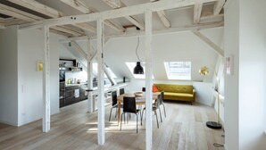 Um amplo loft com vigas de madeira aparentes, área de jantar, sofá amarelo e cozinha ao fundo.