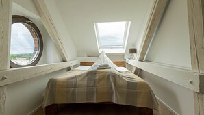 Um quarto de sótão pequeno e triangular com uma cama de solteiro, uma janela redonda e um claraboia.