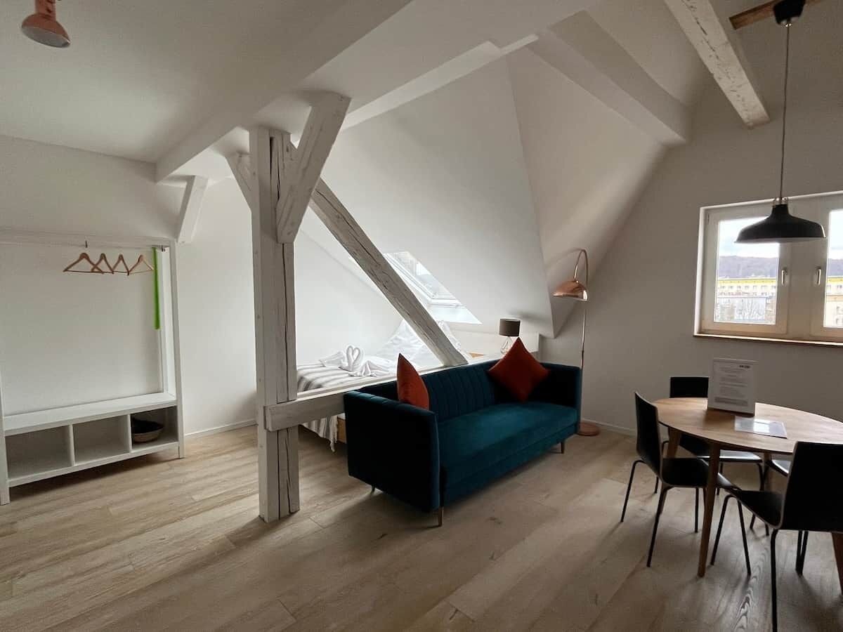 A modern loft bedroom with a sloped ceiling, a bed, a blue sofa with red cushions, a dining table with chairs, and a large window providing a view of the outdoors.