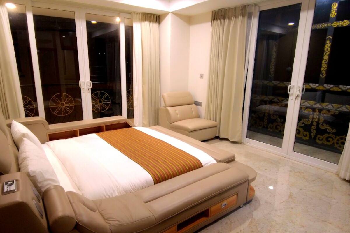 A spacious bedroom with a large bed, a sofa, and a chair. There are sliding glass doors leading to a balcony with a patterned floor.