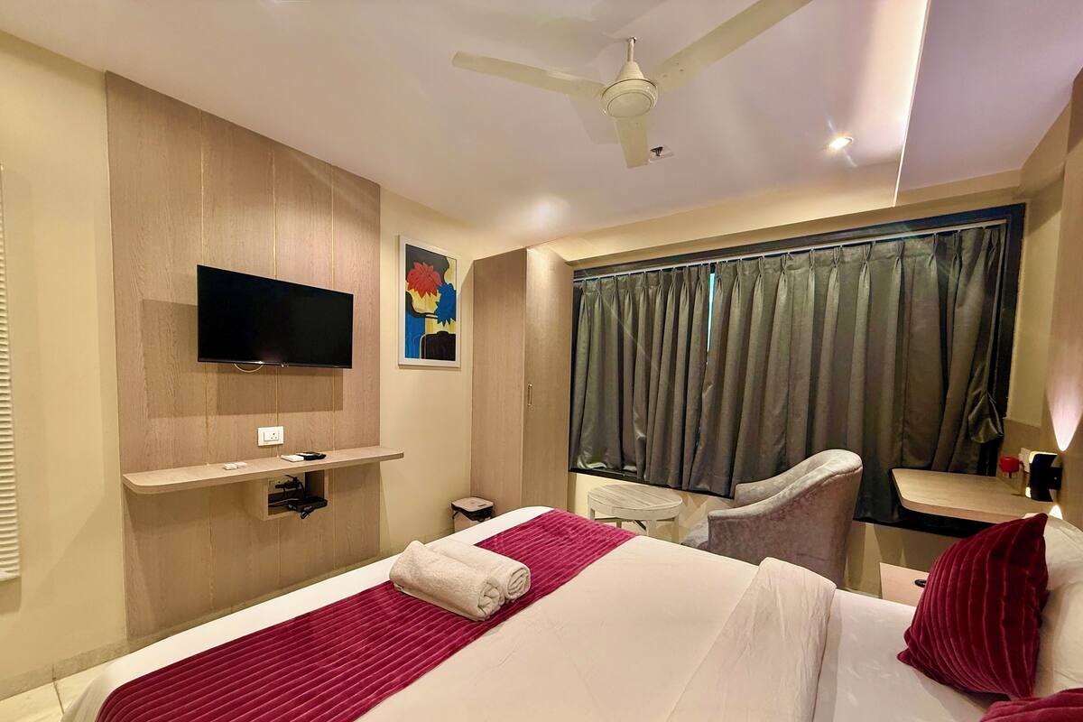 Quarto de hotel com uma cama, televisão, escrivaninha, cadeira e janela com cortinas.