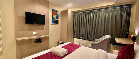 Habitación de hotel con cama, televisor, escritorio, silla y ventana con cortinas.