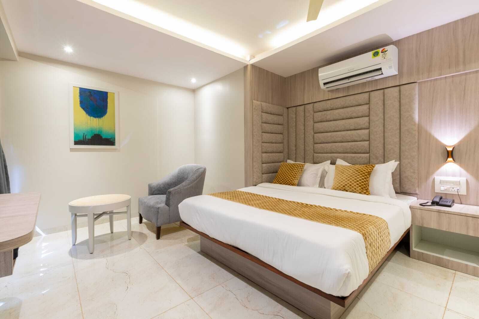 A modern hotel room with a large bed, a chair, a small table, an air conditioning unit, and a wall-mounted lamp.