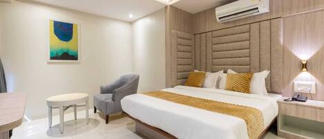A modern hotel room with a large bed, a chair, a small table, an air conditioning unit, and a wall-mounted lamp.