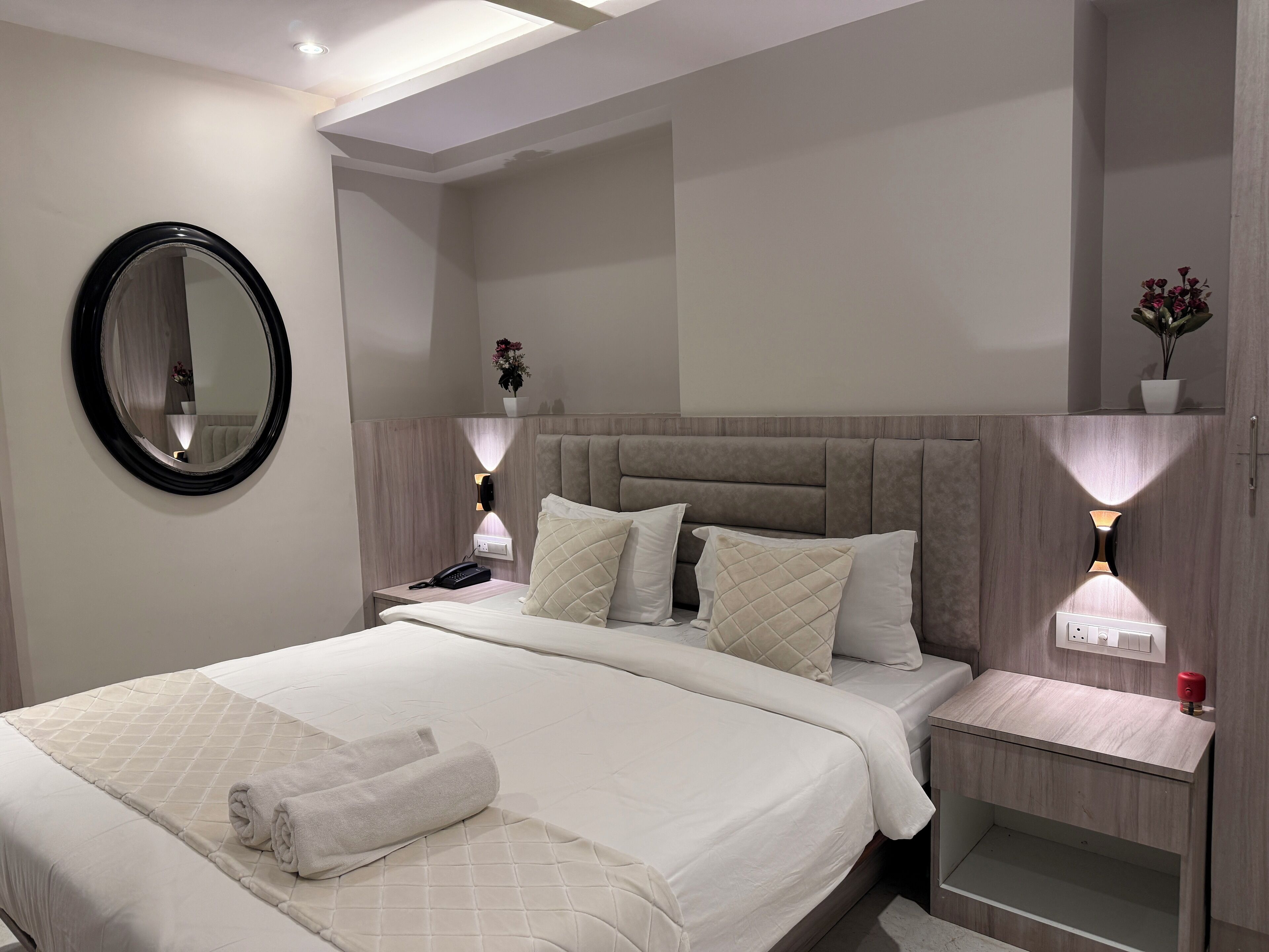 A modern hotel room with a large bed, a bedside table with a lamp, a mirror, and a nightstand with a phone.