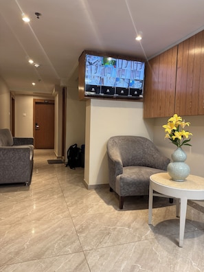 A modern hotel lobby with a flat-screen TV mounted on the wall displaying a map, a grey sofa, a small round table with a vase of yellow flowers, and a wooden door labeled '207'. - kurla suites (Mumbai)