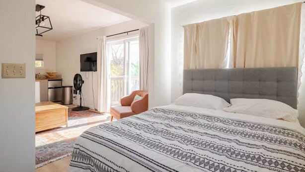 A bedroom with a large bed, a TV mounted on the wall, and a kitchenette in the background.