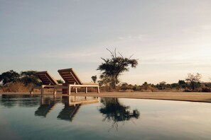 A pool with two lounge chairs, surrounded by dry vegetation and a clear sky.