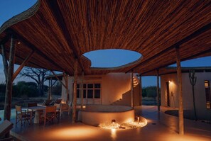 An outdoor dining area with a curved seating arrangement, a central fire feature, and a distinctive wooden ceiling structure.
