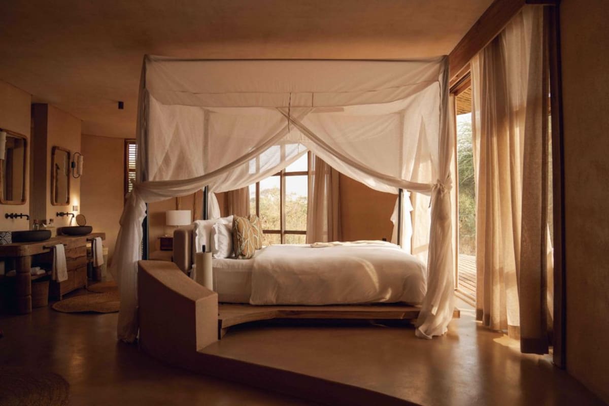 A bedroom with a canopy bed, a sitting area with a sofa, a vanity with a mirror, and a window with curtains.