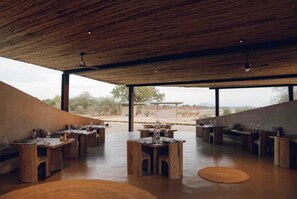 An open-plan dining area with multiple wooden tables and chairs, large windows offering a view of the outdoors.
