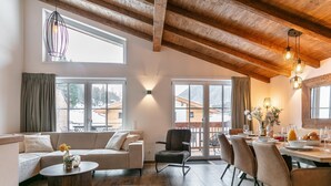 Uma sala de estar ampla com teto de madeira, um conjunto de sofás, uma mesa de jantar com cadeiras e grandes janelas com vista para uma paisagem coberta de neve.