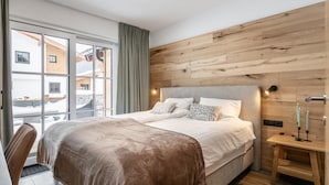 Quarto moderno com uma cama grande, cabeceira de madeira, escrivaninha com abajur e vista para edifícios através de janelas amplas.