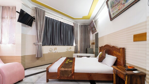 Una habitación de hotel con cama, mesita de noche, silla, televisor montado en la pared y ventana con cortinas.