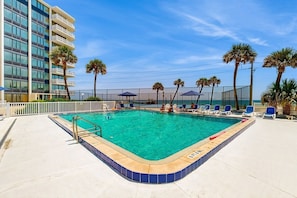 A swimming pool with lounge chairs and palm trees, surrounded by a fence and a building.