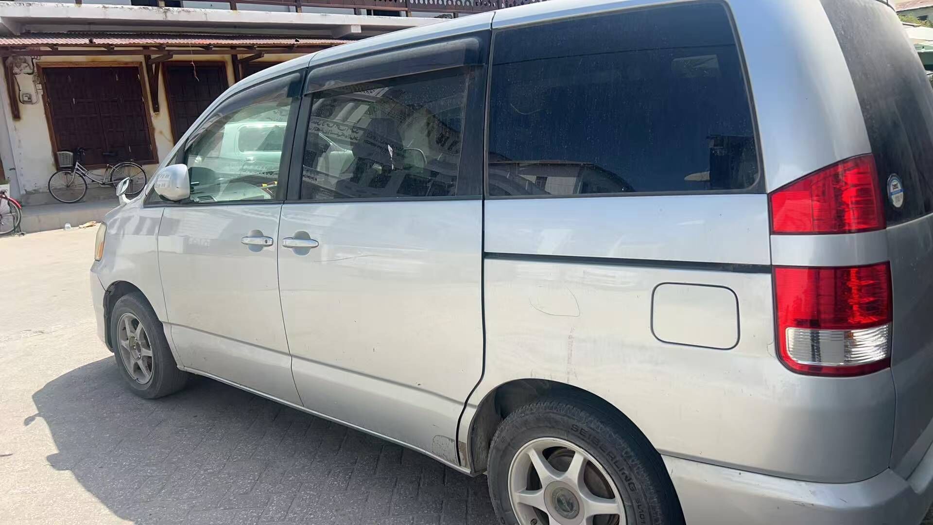 A silver van parked in a lot with a bicycle in the background.